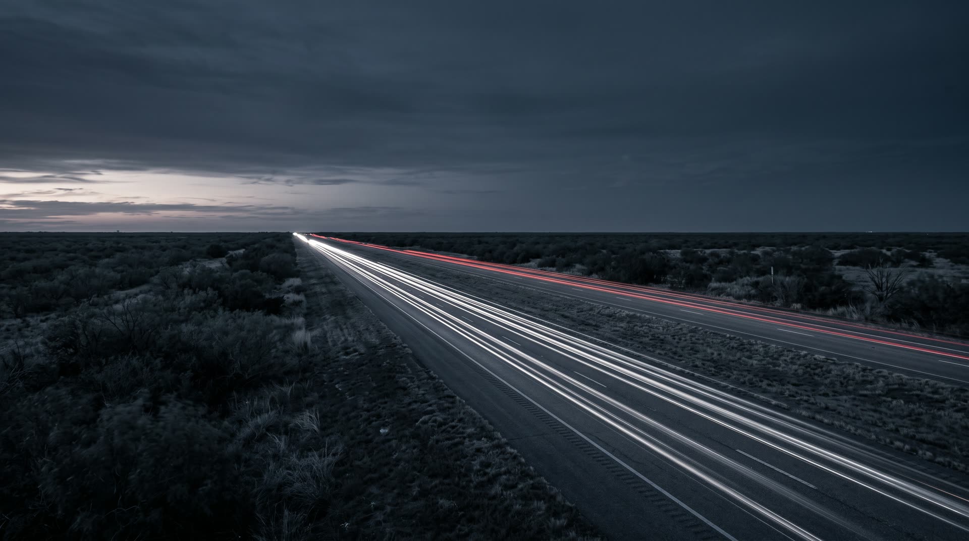 Interstate freight corridor at blue hour with long-exposure truck light trails across a flat Texas horizon
